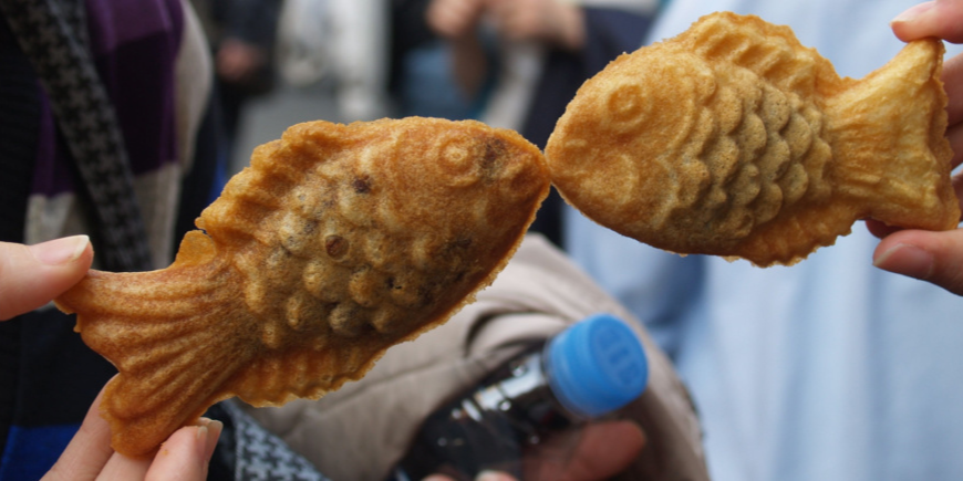 Two people holding up taiyaki cakes, so it looks like the fish are kissing 